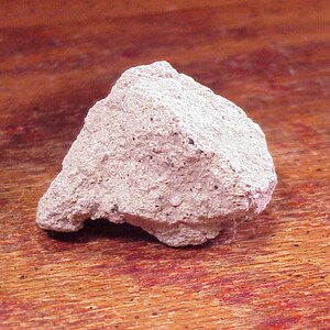 May include: A rough, light beige rock with a porous, uneven texture and small dark speckles throughout its surface. The stone has an irregular, angular shape with a jagged edge and a matte finish, displaying a natural, unpolished appearance.
