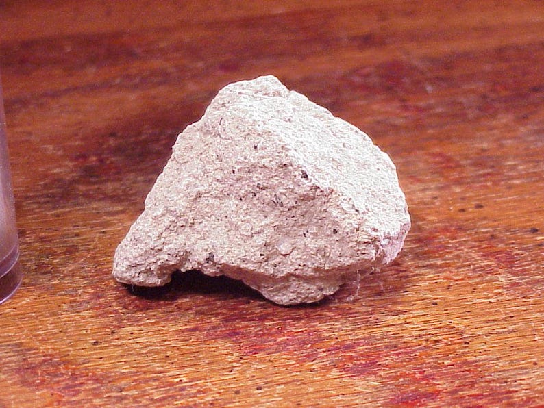 May include: A rough, light beige rock with a porous, uneven texture and small dark speckles throughout its surface. The stone has an irregular, angular shape with a jagged edge and a matte finish, displaying a natural, unpolished appearance.