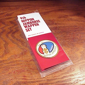 May include: A set of Nippon Jamboree Wappen pins in a clear plastic sleeve. The packaging is red and white with text that reads "9th Nippon Jamboree Wappen Set" and "Boy Scouts of Nippon". A round pin with a blue, red, and gold design is visible.