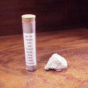 May include: A clear cylindrical plastic vial with a cork stopper containing grey volcanic ash, labeled Mt St Helens Ash and Pumice Stone, May 18th, 1980 Eruption, positioned next to a single, rough, light grey piece of volcanic pumice stone.
