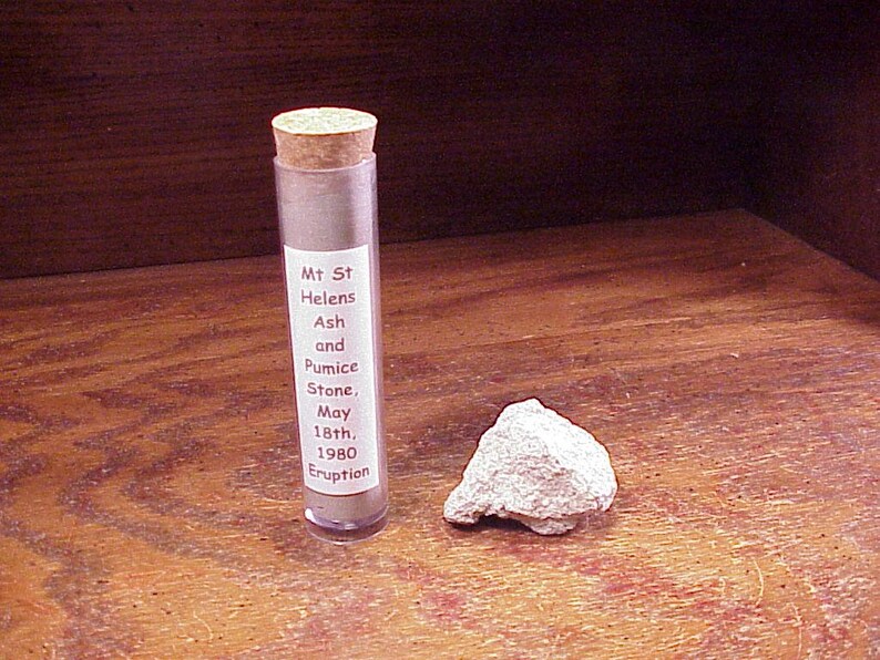 May include: A clear cylindrical plastic vial with a cork stopper containing grey volcanic ash, labeled Mt St Helens Ash and Pumice Stone, May 18th, 1980 Eruption, positioned next to a single, rough, light grey piece of volcanic pumice stone.