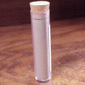 May include: A clear, cylindrical plastic tube containing a rolled-up piece of paper, sealed at the top with a light brown, textured cork stopper. The tube is transparent, revealing the light-colored paper inside, and features a small white label.
