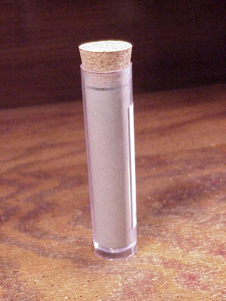 May include: A clear, cylindrical plastic tube containing a rolled-up piece of paper, sealed at the top with a light brown, textured cork stopper. The tube is transparent, revealing the light-colored paper inside, and features a small white label.