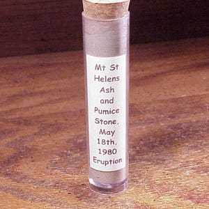May include: A clear cylindrical plastic vial filled with gray volcanic ash and pumice stone, sealed with a tan cork stopper. A white rectangular label on the front reads: Mt St Helens Ash and Pumice Stone, May 18th, 1980 Eruption in dark text.
