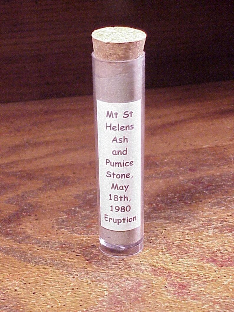 May include: A clear cylindrical plastic vial filled with gray volcanic ash and pumice stone, sealed with a tan cork stopper. A white rectangular label on the front reads: Mt St Helens Ash and Pumice Stone, May 18th, 1980 Eruption in dark text.