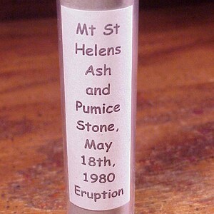 May include: A clear cylindrical tube containing grey volcanic ash and pumice stone. A white label on the front features the text: Mt St Helens Ash and Pumice Stone, May 18th, 1980 Eruption. The container is upright and filled with granular material.
