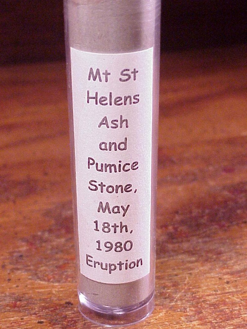 May include: A clear cylindrical tube containing grey volcanic ash and pumice stone. A white label on the front features the text: Mt St Helens Ash and Pumice Stone, May 18th, 1980 Eruption. The container is upright and filled with granular material.