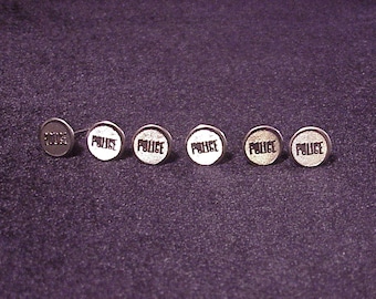 May include: Six silver-colored metal buttons with the word "POLICE" embossed on each. The buttons are arranged in a row on a dark purple fabric background.