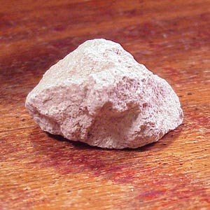 May include: A single, irregularly shaped rock with a rough, porous texture and a pale, dusty pinkish-beige color. The stone features sharp, jagged edges and a matte surface, displaying natural variations in its mineral composition and structure.