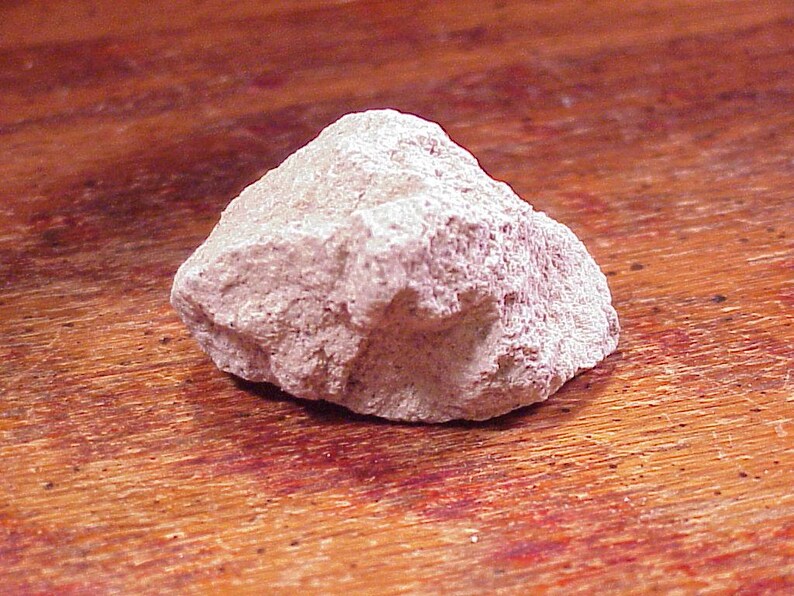 May include: A single, irregularly shaped rock with a rough, porous texture and a pale, dusty pinkish-beige color. The stone features sharp, jagged edges and a matte surface, displaying natural variations in its mineral composition and structure.