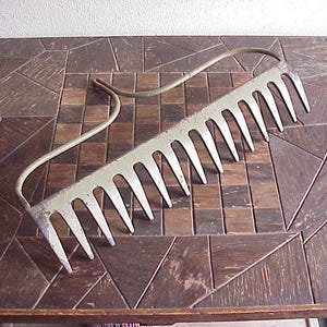 May include: A vintage metal garden rake with a curved handle and multiple tines. The rake is painted a light green color and rests on a dark brown, geometric patterned surface.