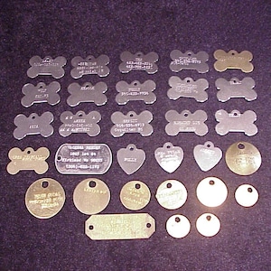 May include: A collection of 25 silver and gold dog tags. The tags are in various shapes, including bone, heart, and circle. Some tags have engraved text, including names, phone numbers, and addresses.