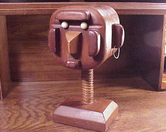 May include: A wooden sculpture of a face with a spring-loaded base. The face has a brown wooden body with white eyes and a brown wooden nose. The sculpture is on a wooden base.