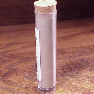 May include: A clear cylindrical plastic tube filled with a light tan powder, sealed with a round cork stopper. A white rectangular label is attached to the side of the tube, featuring partially visible black printed text in a vertical orientation.