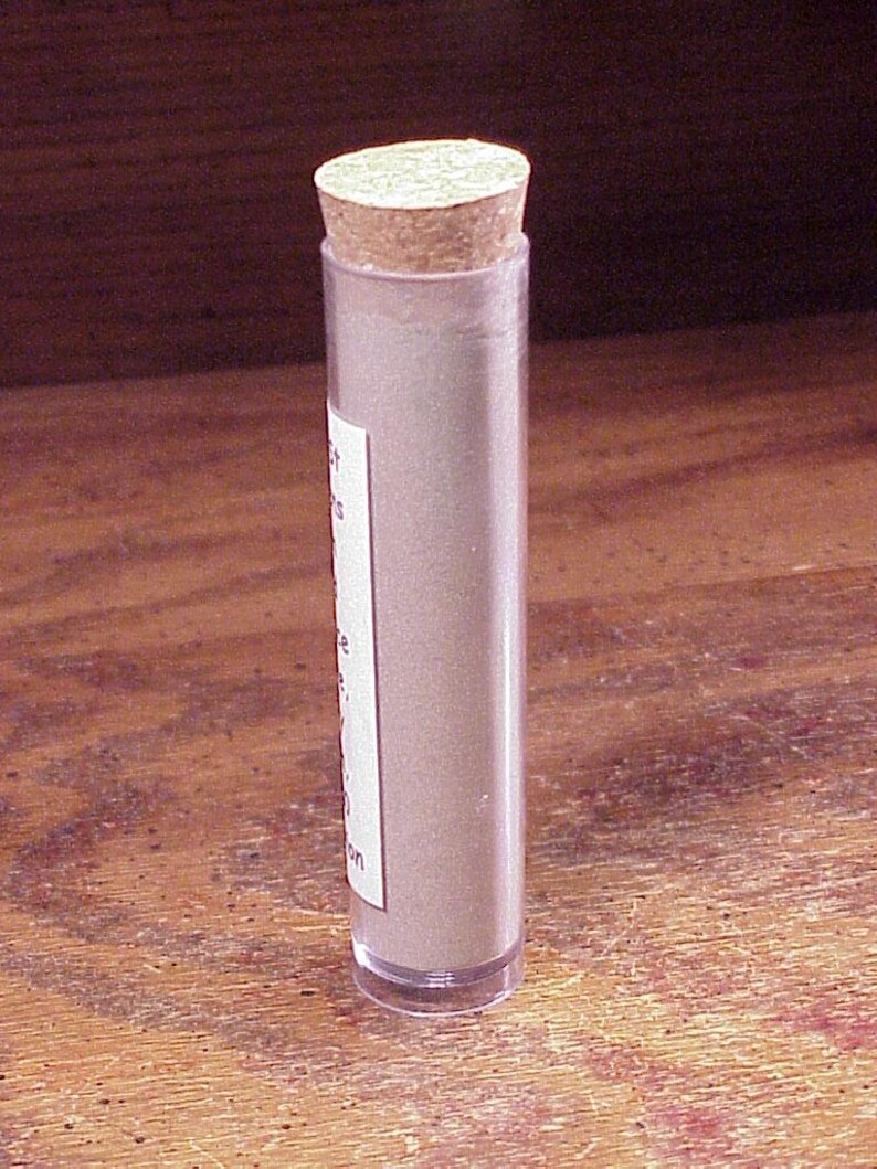 May include: A clear cylindrical plastic tube filled with a light tan powder, sealed with a round cork stopper. A white rectangular label is attached to the side of the tube, featuring partially visible black printed text in a vertical orientation.