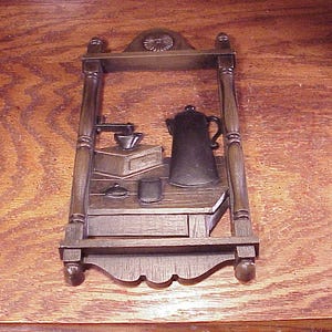 May include: A brown, wall-mounted shelf with a coffee-themed design. It features a coffee pot, grinder, cup, and other accessories. The shelf has a decorative top and bottom, and is made of wood.