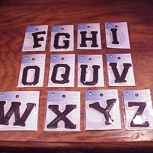 May include: A set of 12 black iron-on letters in uppercase, including the letters F, G, H, I, O, Q, U, V, W, X, Y, and Z. The letters are packaged individually on white cards with blue borders.