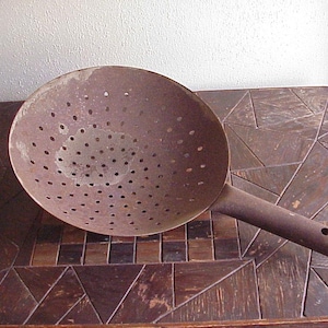 May include: A rusty, round metal colander with a long handle and numerous small holes. The kitchen utensil is designed for draining liquids from food. The colander is resting on a wooden table.
