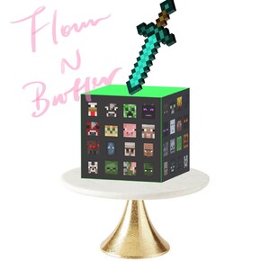 May include: A green Minecraft cake with a diamond sword on top. The cake is decorated with Minecraft character faces. The cake is on a white cake stand with a gold base.