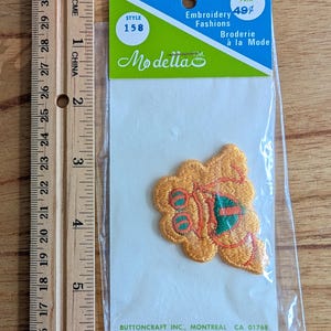 Può includere: Una toppa ricamata arancione e verde in una confezione di plastica trasparente. La toppa ha la forma di un fiore o di una foglia. La confezione riporta la scritta "Embroidery Fashions" e "Modelia". Un righello è accanto alla confezione.