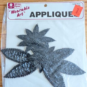 Peut inclure: Applique noire à paillettes et perles dans un emballage plastique transparent. L'applique a la forme d'une fleur en forme d'éclat d'étoile. L'emballage porte les mots "Wearable Art" et "APPLIQUE".