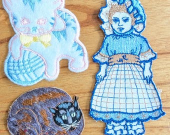 May include: Three embroidered patches: a light blue cat with a yellow bow, a sleeping calico cat, and a blue doll in a dress. The patches are on a wooden surface. The cat patches have pink accents.