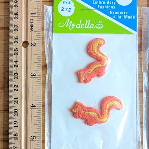 Peut inclure: Deux écussons brodés de l'écureuil orange et jaune, emballés dans une pochette en plastique transparent. L'emballage comprend le texte "Modella" et "Embroidery Fashions". Une règle en bois est sur le côté gauche de l'image.