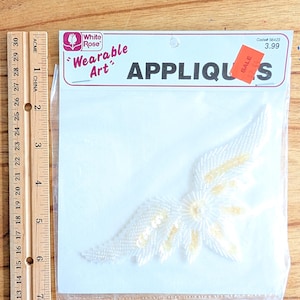 May include: A white and gold sequined applique in a clear plastic package. The applique is in the shape of a wing or feather. The package has the words "Wearable Art" and "APPLIQUES" printed on it.