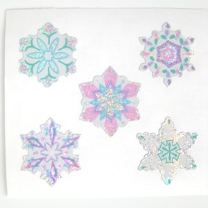 May include: Five iridescent snowflake stickers with a white background. The snowflakes are in various shades of pink, blue, and green.