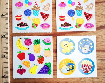 Può includere: Quattro fogli di adesivi con vari disegni di cibo e frutta. Il primo foglio ha un hot dog, un hamburger, un cono gelato, un cupcake, popcorn, un lecca-lecca, patatine fritte, pizza e una ciambella. Il secondo foglio ha gli stessi disegni del primo foglio. Il terzo foglio ha una banana, un ananas, un'anguria, una mela, una prugna, una fragola, un'arancia, una pera e dell'uva. Il quarto foglio ha quattro adesivi rotondi con popcorn e uno schermo televisivo.