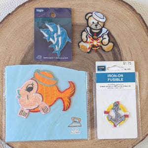May include: A collection of iron-on patches featuring nautical and marine themes. Includes a dolphin design, a teddy bear in a sailor outfit, a cartoon fish, and an anchor within a life preserver.