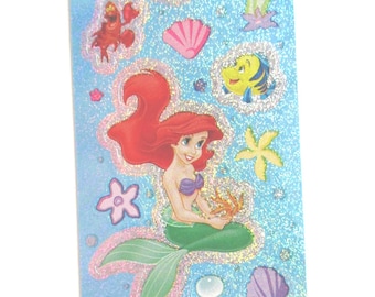 Puede incluir: Una hoja de pegatinas con Ariel, la Sirenita, en varias poses. Las pegatinas están sobre un fondo azul con purpurina e incluyen otros personajes como Sebastián y Flounder. La hoja está etiquetada con el copyright de Disney.