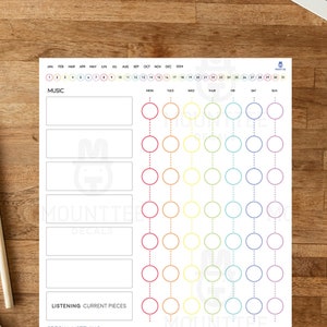 Puede incluir: Un rastreador de escucha de música blanco con un degradado arcoíris de círculos, una sección para notas y el texto "MOUNTTEE". Dos lápices y una pequeña planta en maceta se encuentran sobre una superficie de madera junto a una computadora portátil.