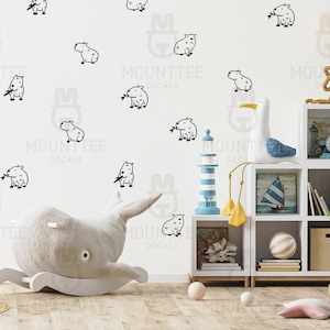 Peut inclure: Un mur blanc avec des illustrations d'animaux en noir et blanc, dont un cheval à bascule en forme de baleine, une mouette et un phare. Une bibliothèque blanche avec des étagères remplies de livres, un voilier, une coquille de mer et une tour de blocs en bois. Le texte "MOUNTTEE DECALS" apparaît plusieurs fois sur le mur.