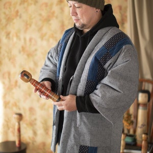 Könnte beinhalten: Eine Person trägt eine graue und blaue Patchworkjacke, eine grüne Mütze und eine blaue Jeans und hält eine Holzpuppe. Die Puppe ist mit roten, gelben und schwarzen Streifen bemalt.