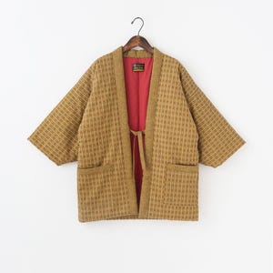 May include: A tan kimono-style jacket with a vertical striped pattern. The jacket has wide sleeves, two front pockets, and a red interior lining. It is hanging on a wooden hanger against a white background.
