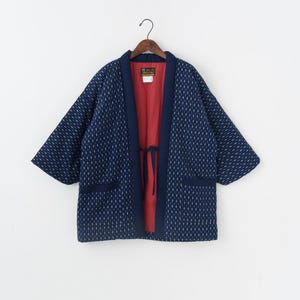 Puede incluir: Chaqueta azul marino estilo kimono con un estampado de puntos blancos. La chaqueta tiene un interior rojo y ribetes azul marino. Cuenta con cierre de lazo y dos bolsillos delanteros. La chaqueta está colgada de una percha de madera.