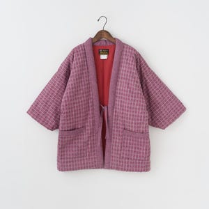 Puede incluir: Chaqueta estilo kimono de color malva con un patrón de rayas verticales. La chaqueta tiene mangas cortas, dos bolsillos delanteros y un forro interior rojo. Cuelga de una percha de madera.