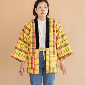 Könnte beinhalten: Gelb karierte Jacke mit roten und blauen Akzenten und einem schwarzen Kragen. Die Jacke hat einen Bindverschluss und wird über einem weißen Hemd und einer blauen Jeans getragen. Die Jacke hat eine lockere Passform und dreiviertellange Ärmel.
