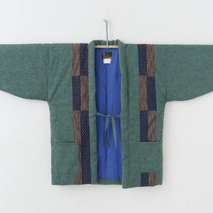 Op de afbeelding: Een groene gewatteerde jas met een blauwe binnenkant, hangend aan een houten roede. De jas heeft decoratieve panelen van bruine en marineblauwe stof. Het heeft een striksluiting en twee voorzakken.