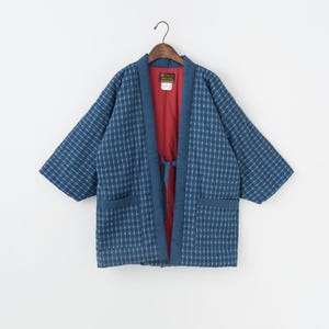 Puede incluir: Chaqueta azul y blanca estampada con interior rojo. La chaqueta tiene un diseño envolvente con cinturón a juego, dos bolsillos delanteros y mangas tres cuartos. Cuelga de una percha de madera.