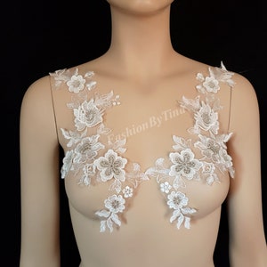 May include: Two white floral lace appliques with a beaded design. The appliques are on a white mannequin bust.
