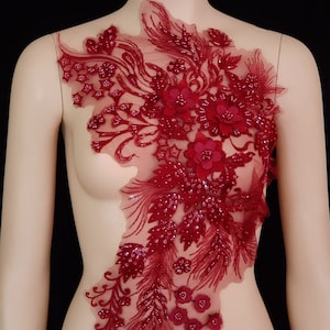 May include: A burgundy lace applique with beaded embellishments and floral designs. The applique features a large central flower with smaller flowers and leaves surrounding it.