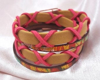 May include: A multi-layered bracelet with a tan leather base and pink suede accents. The leather is woven in a criss-cross pattern and the bracelet has a colorful, abstract print.