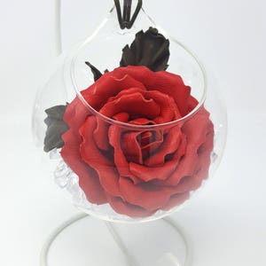 May include: A single red leather rose is displayed in a clear glass orb. The rose is surrounded by white crystals and has a single dark green leather leaf.