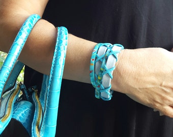 May include: A turquoise crocodile embossed leather handbag with a gold zipper. A woman's wrist is wearing a stack of turquoise and white leather bracelets with red and yellow accents.