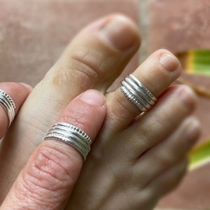 May include: Three silver toe rings with a textured design. The rings are stacked on the toe of a person's foot.