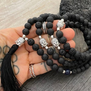 Puede incluir: Cuentas de mala de piedra de lava negra con acentos de cabeza de Buda plateados y un borla negra. Las cuentas de mala están enhebradas en un cordón negro y tienen una cuenta de piedra azul.