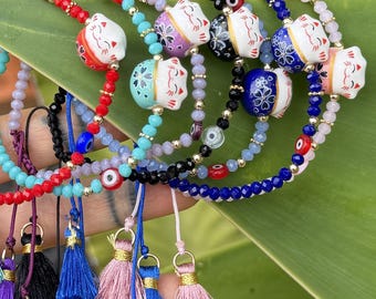 Op de afbeelding: Een collectie verstelbare armbanden met keramische maneki-neko kattenkralen, kleurrijke gefacetteerde glaskralen en kleine boze oog bedels. Elke armband is afgewerkt met een bungelend stoffen kwastje in diverse kleuren zoals rood, blauw en roze.