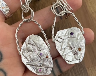 May include: Two silver necklaces with geometric pendants featuring colorful gemstones. The pendants are shaped like a jagged rock and have a textured surface.
